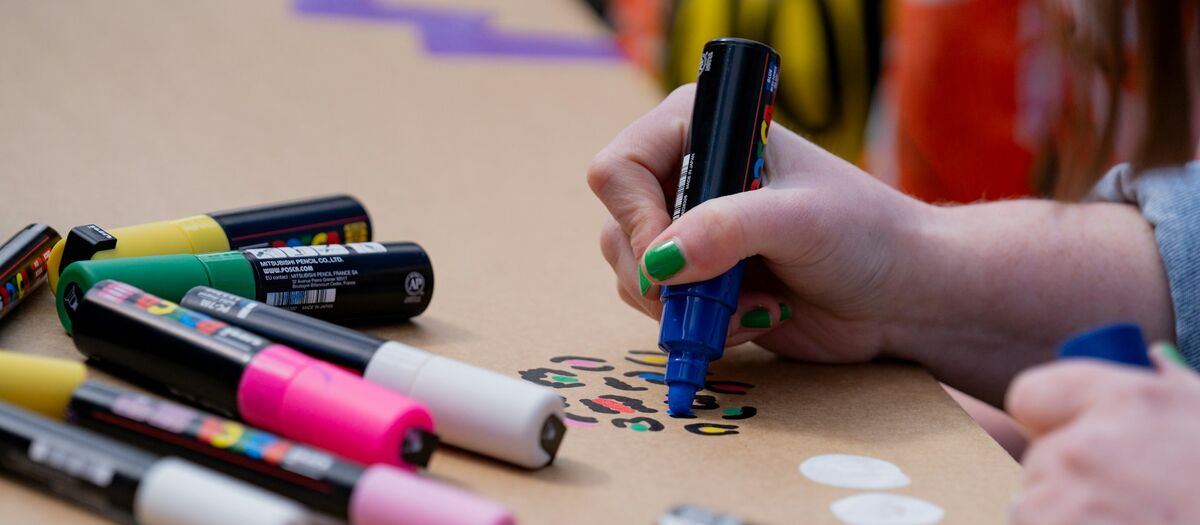 Hand mit grüner Nagellackierung hält einen blauen Marker und malt auf braunes Papier, daneben liegen mehrere bunte Marker.